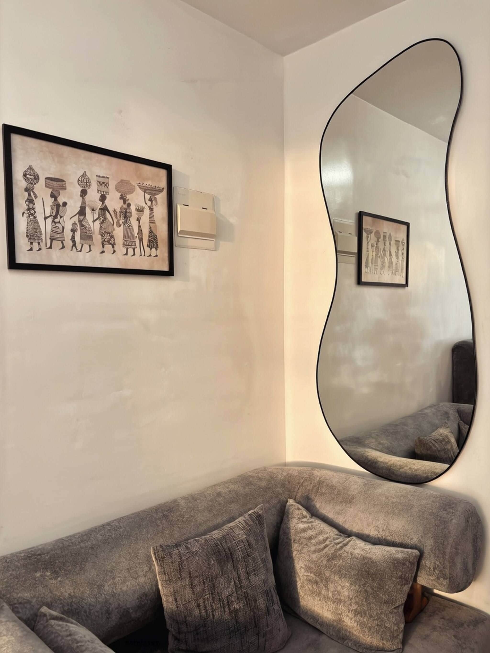 Una sala de estar moderna con un sofá gris, un espejo abstracto grande y dos obras de arte enmarcadas en la pared.