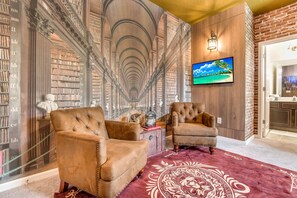 Eine Bibliothek mit Bücherregalen, zwei Ledersesseln, einem roten Teppich mit Muster und einem an der Wand montierten Fernseher.