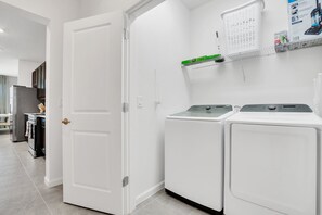A laundry room with two white washing machines, a shelf with cleaning supplies, and a door leading to another room. - Grand 15BR Kissimmee Estate 30 Guests (Kissimmee)