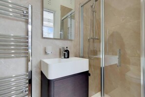 A modern bathroom with a glass shower enclosure, a white sink, and a dark cabinet. - DG1 - Family Friendly 2-Bed with Garden (London)