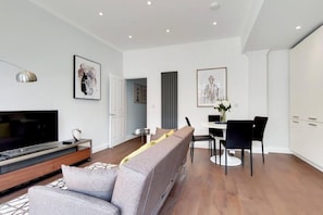 Ein modernes Wohnzimmer mit Flachbildfernseher, grauem Sofa, Esstisch mit Stühlen und Kunstwerken an den Wänden.