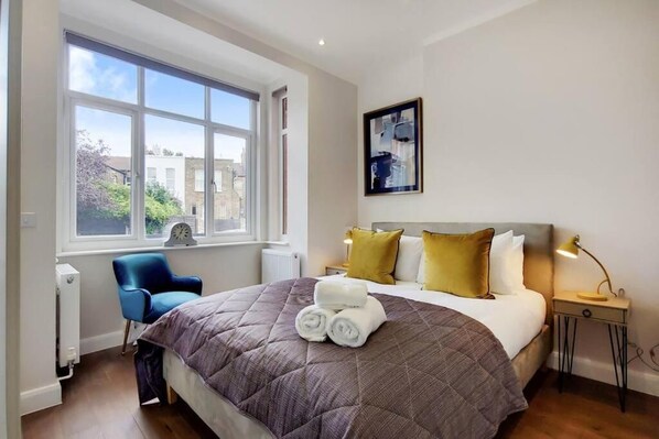 A hotel room with a large window, a bed with a quilt, a chair, a bedside table with a lamp, and a wall-mounted picture. - DG1 - Family Friendly 2-Bed with Garden (London)