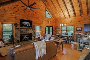 Ein Interieur einer Holzhütte mit Kamin aus Stein, Ledersofa, Esstisch und einem an der Wand befestigten Fernseher.