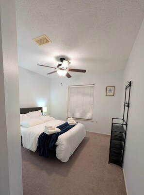Une chambre à coucher avec un lit, un ventilateur de plafond, une fenêtre avec des stores, une étagère fixée au mur et un tableau encadré.