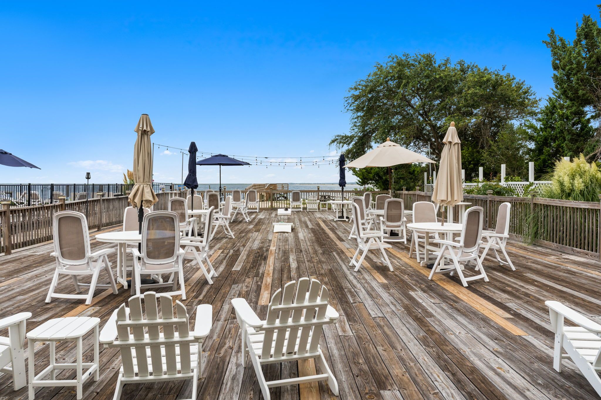 A wooden deck with white chairs and tables, string lights, and umbrellas.