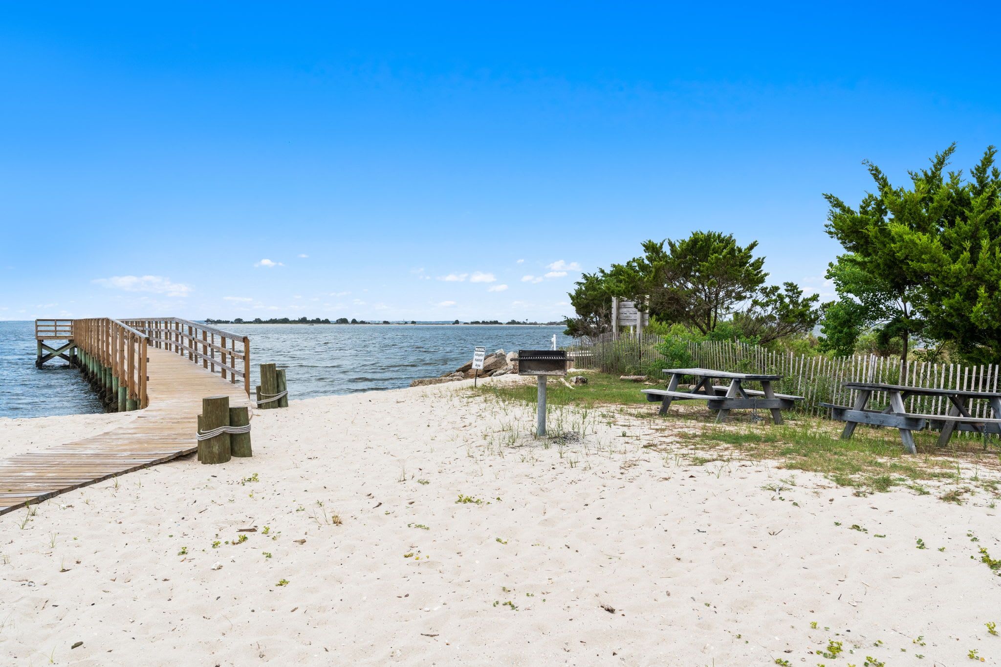 A sandy beach with a wooden pier extending into the water, a picnic table, and a signpost.