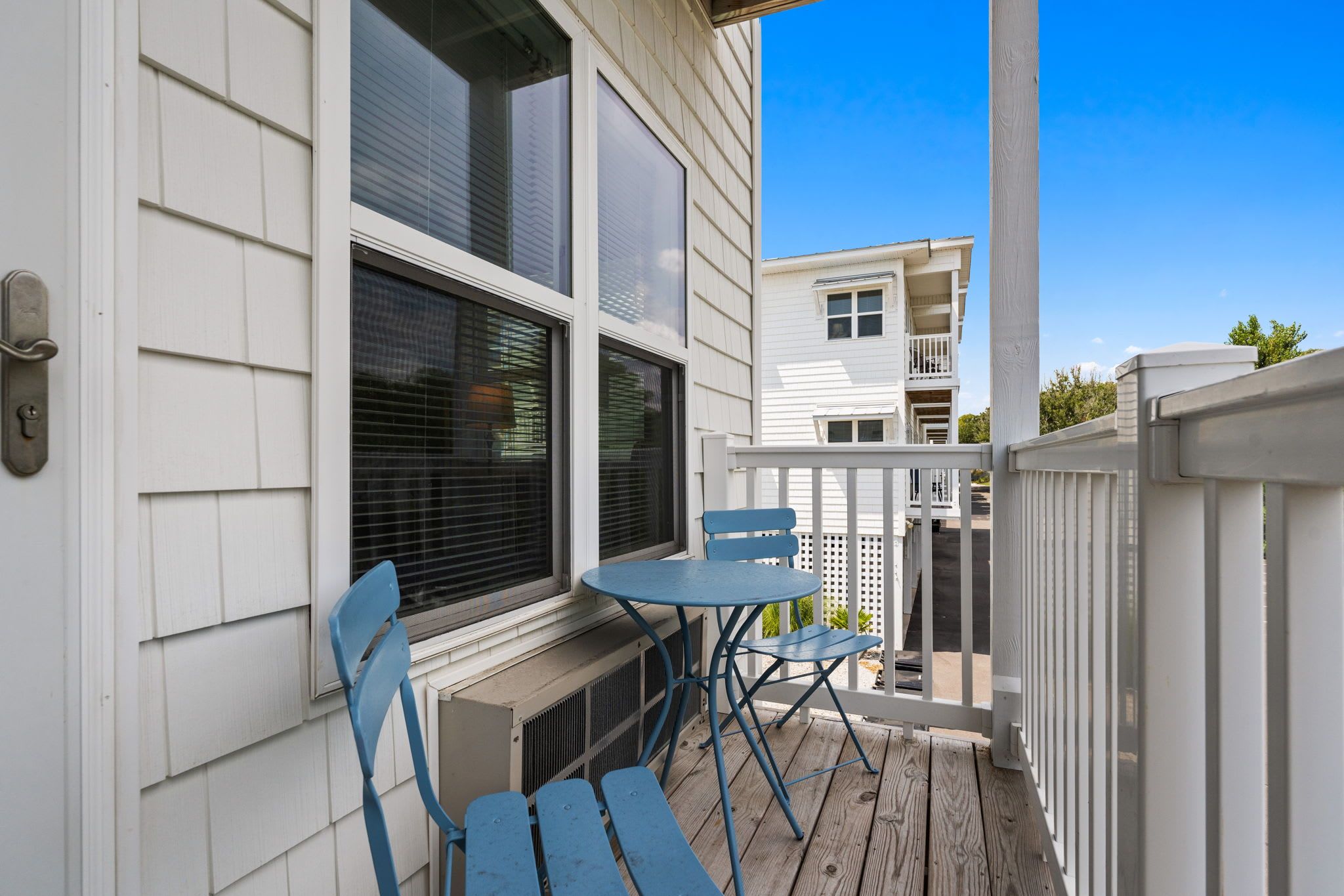 A balcony with a table and chairs, a window with blinds, and a white building in the background.