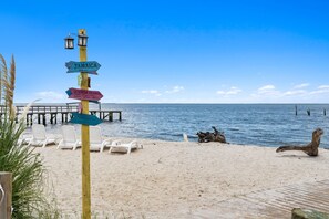Una playa con un cartel de madera, sillones blancos para tomar el sol y un muelle que se adentra en el agua.
