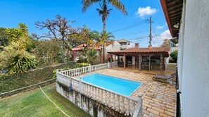 A swimming pool with a white balustrade, surrounded by a garden with various plants and a patio area with outdoor furniture. - Casa com Piscina e Sauna no Granja Vista Alegre (Contagem)