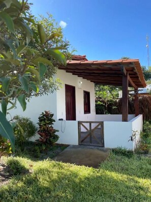 Un pequeño edificio blanco con una pérgola de madera, un portón de madera y un jardín bien cuidado.