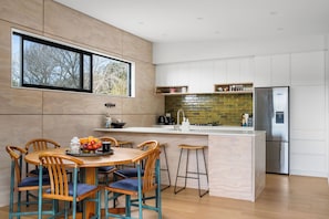 A modern kitchen with a central island, bar stools, and a dining table with chairs. - The Cove Escape - Cooks Beach Holiday Home (Cooks Beach)