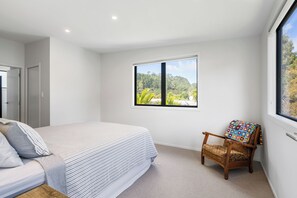 A bedroom with a bed, a chair, a window with a view of a forest, and a door. - The Cove Escape - Cooks Beach Holiday Home (Cooks Beach)