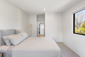 A bedroom with a bed, a window with a view of trees, and a door leading to another room. - The Cove Escape - Cooks Beach Holiday Home (Cooks Beach)