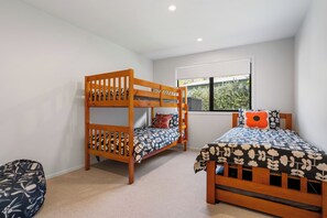 A room with bunk beds and a single bed, carpeted floor, and a window with a view of greenery. - The Cove Escape - Cooks Beach Holiday Home (Cooks Beach)