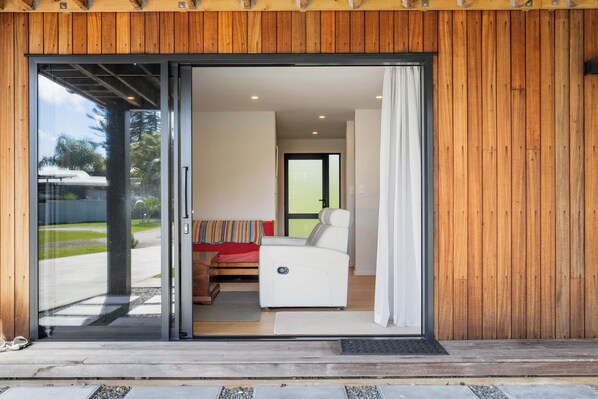 A modern interior with a sliding glass door leading to a room with a bed, a white chair, and a small table. - The Cove Escape - Cooks Beach Holiday Home (Cooks Beach)