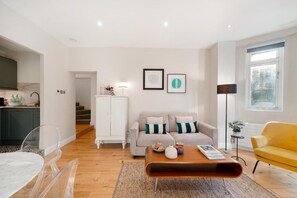 A modern living room with a sofa, coffee table, and a yellow armchair. There is a kitchen area with cabinets and a countertop visible in the background. - HR2 - Charming 1-Bed near Holland Park (London)
