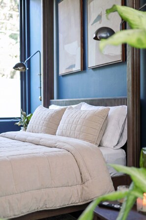 A bed with a quilted white comforter and pillows, a bedside lamp, and a window with a view of greenery. - Luxury SoCo Haven w/ 6Sleep, Hot Tub & Gym (Austin)