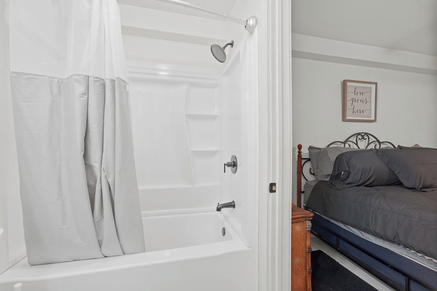 A bathroom with a bathtub, a shower, and a toilet. There is a bed with a headboard and pillows in the adjacent room.