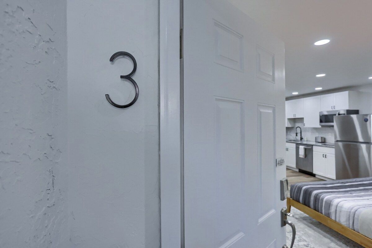 A modern bedroom with a bed, a door with the number 3, and a view into a kitchen area with appliances.