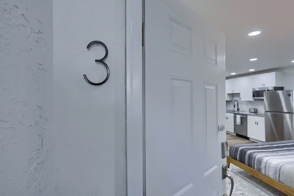 Une chambre moderne avec un lit, une porte portant le numéro 3 et une vue sur un coin cuisine équipé d’appareils électroménagers.