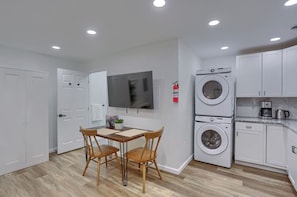 Une cuisine compacte avec des meubles blancs, un lave-linge encastré, une table à manger avec des chaises et un téléviseur fixé au mur.