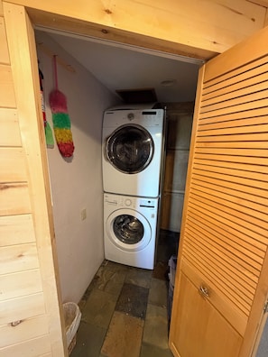 A laundry room with a stacked washer and dryer, a broom, and a trash can.