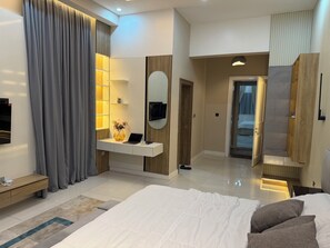 A modern hotel room with a large bed, a bedside table, a desk with a laptop, a mirror, and a wardrobe. - Smart Luxury 2BR | Starlink • PS5 • 24/7 Power • Modern Living (Abuja)