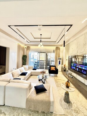 A modern living room with a sectional sofa, a black armchair, a coffee table, and a flat-screen TV mounted on the wall. - Smart Luxury 2BR | Starlink • PS5 • 24/7 Power • Modern Living (Abuja)