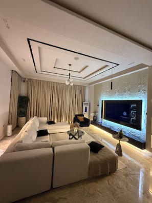 A modern living room with a sectional sofa, a flat-screen TV, and a decorative wall. - Smart Luxury 2BR | Starlink • PS5 • 24/7 Power • Modern Living (Abuja)