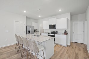 A modern kitchen with white cabinetry, a stainless steel refrigerator, a microwave, and a built-in oven. There are three bar stools and a large island with a sink.