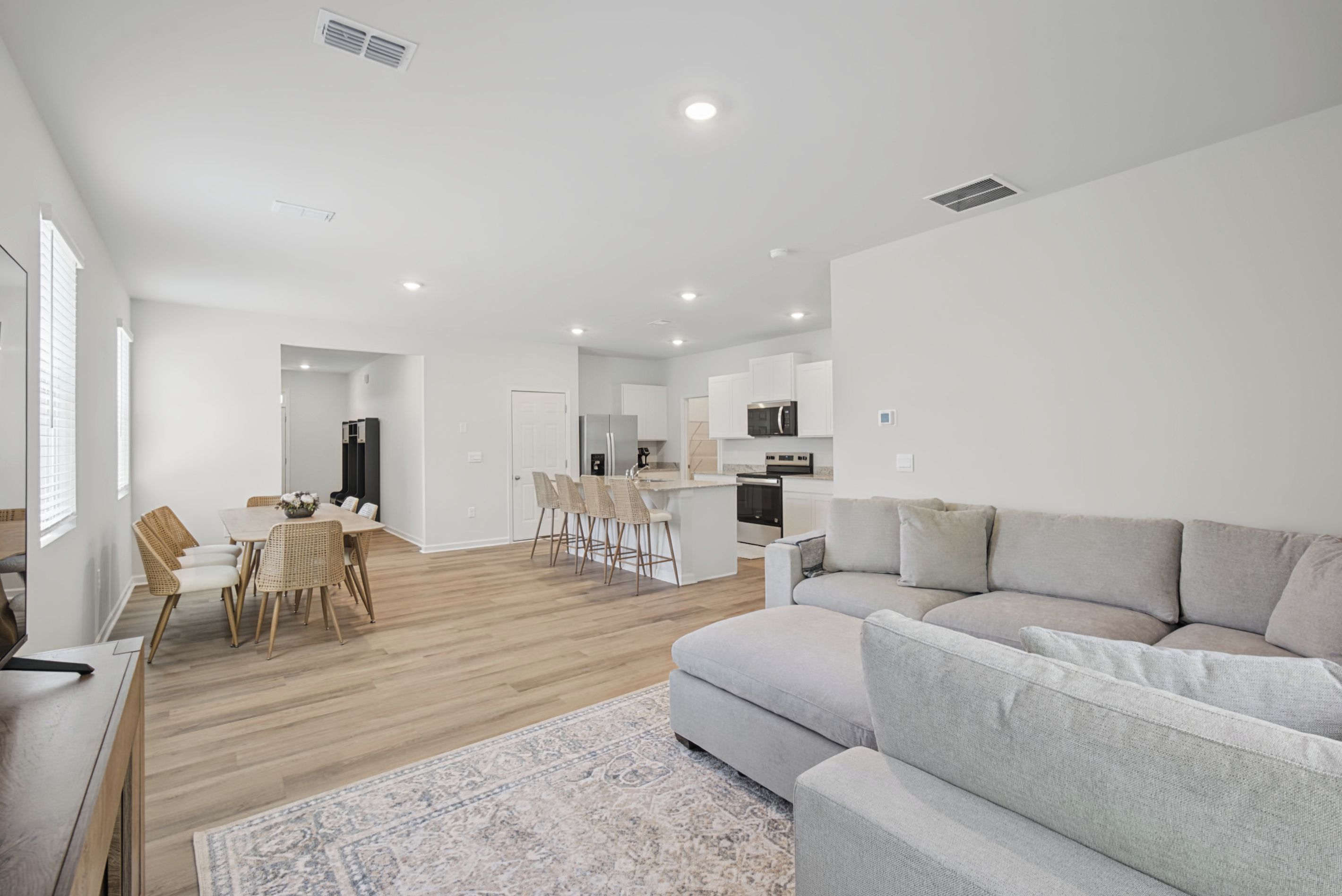 A modern living room with a grey sofa, a dining area with wicker chairs, and a kitchen with white cabinets and a microwave.
