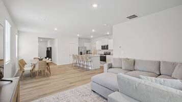 A modern living room with a grey sofa, a dining area with wicker chairs, and a kitchen with white cabinets and a microwave.