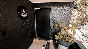 Eine schwarze Tür mit einem runden Schild darüber, ein Blumentopf mit gelben Blättern und eine strukturierte Wand.
