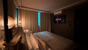 Habitación de hotel con cama, lámpara de noche, televisor de pared mostrando 'NETFLIX' y ventana con persianas verticales.
