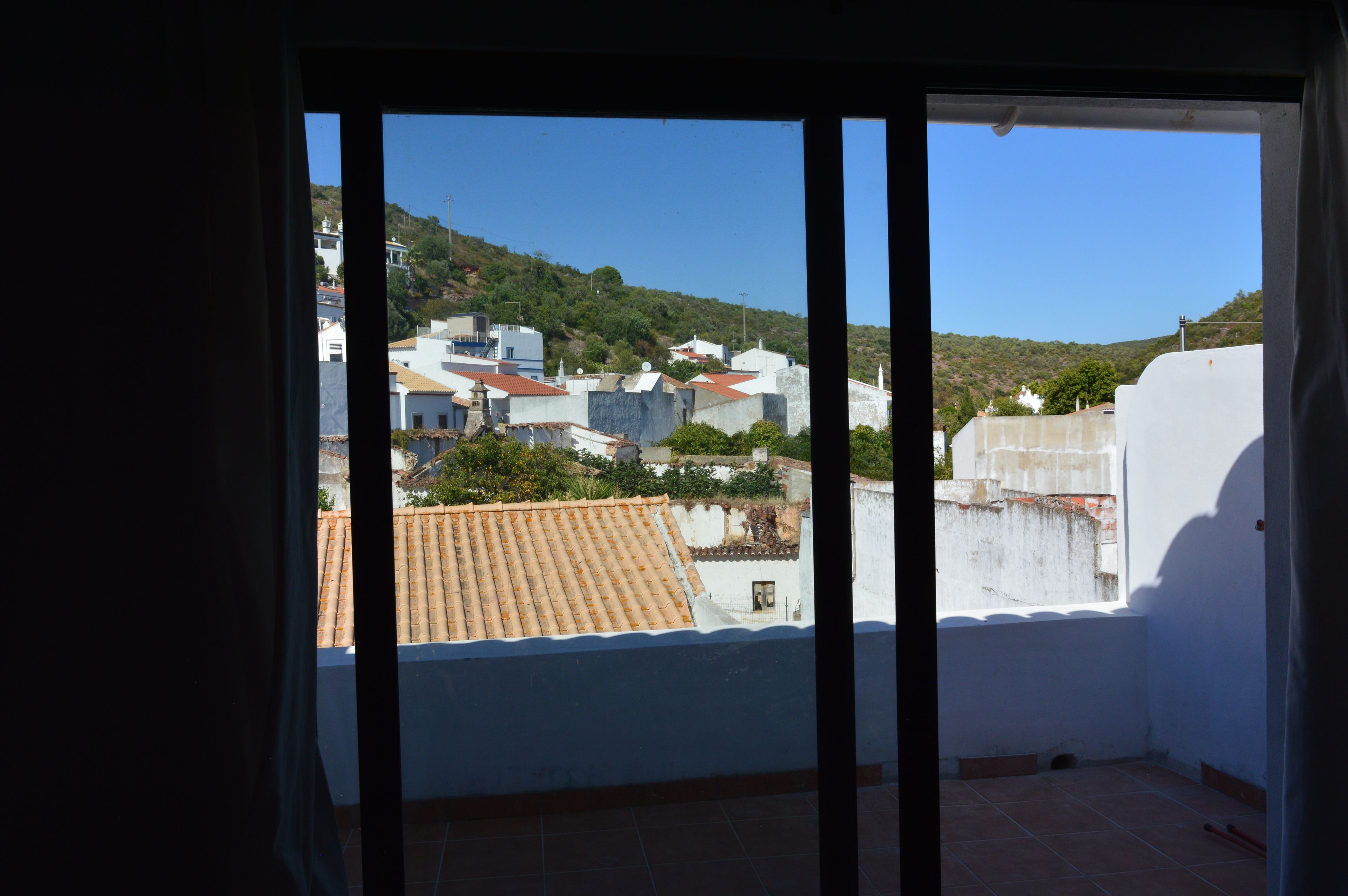 A view from a balcony with a clear sky, a hillside in the background, and buildings with red-tiled roofs.