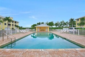 Una piscina con un indicador de profundidad de 6 pies, rodeada por un solárium de ladrillo, con un pequeño edificio al fondo y palmeras.