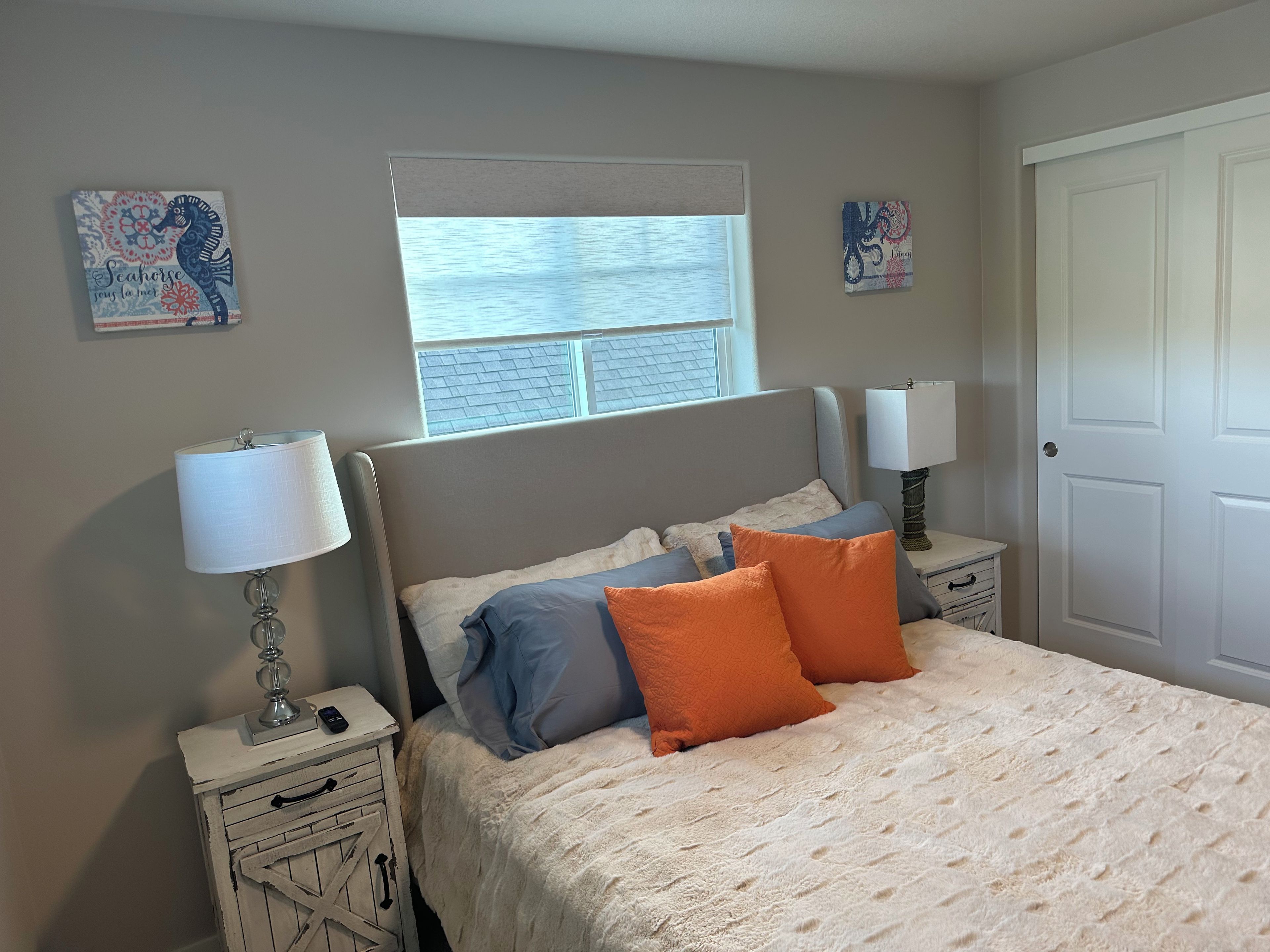 Une chambre à coucher comprenant un lit, deux tables de chevet, une commode, une fenêtre avec des stores et deux œuvres d’art murales.