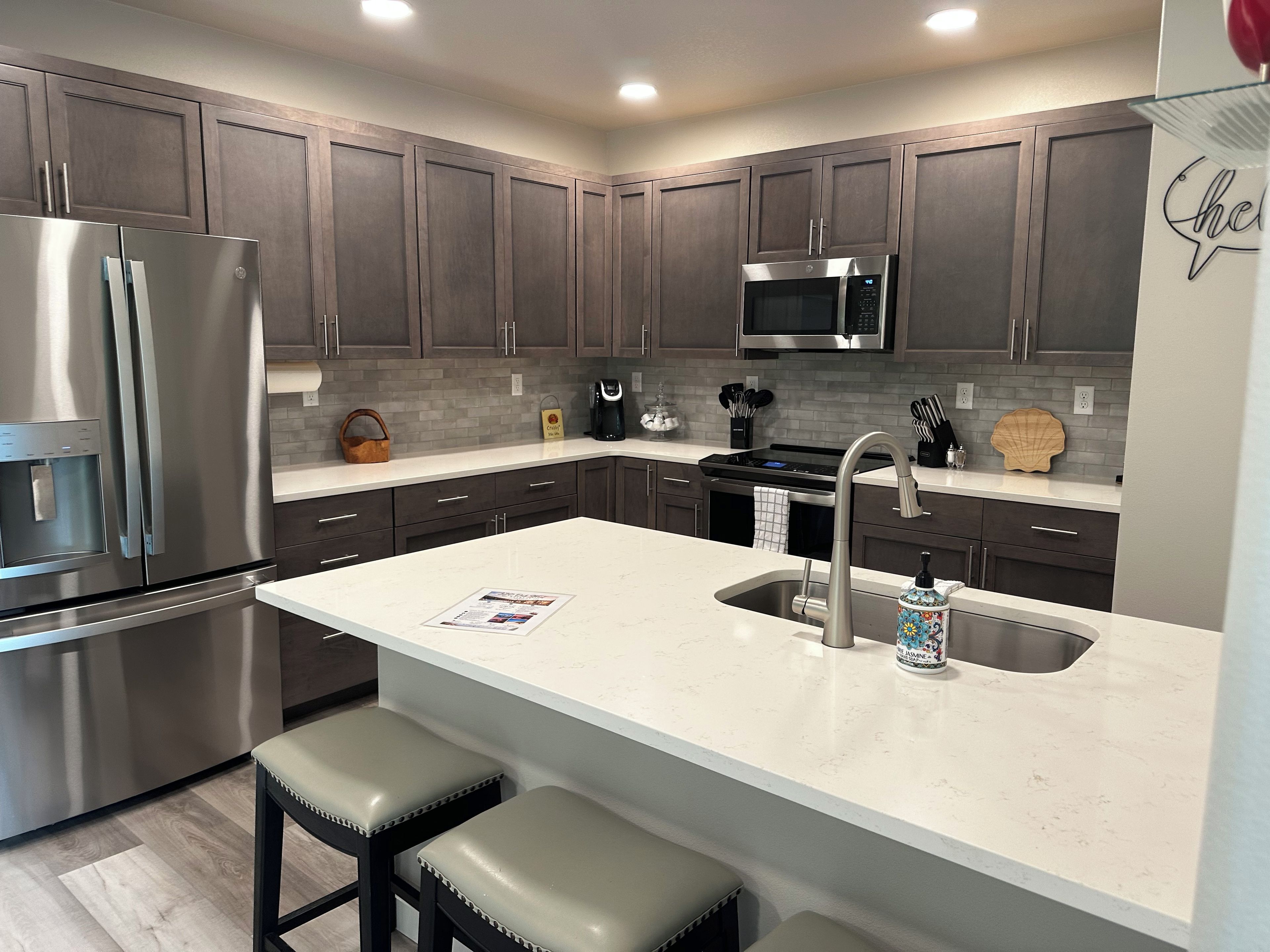 A modern kitchen with stainless steel appliances, dark wood cabinets, a white countertop, and bar stools.