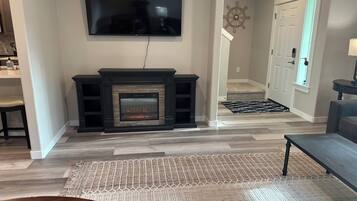A living room with a fireplace, a television mounted on the wall, and a hallway leading to a door.