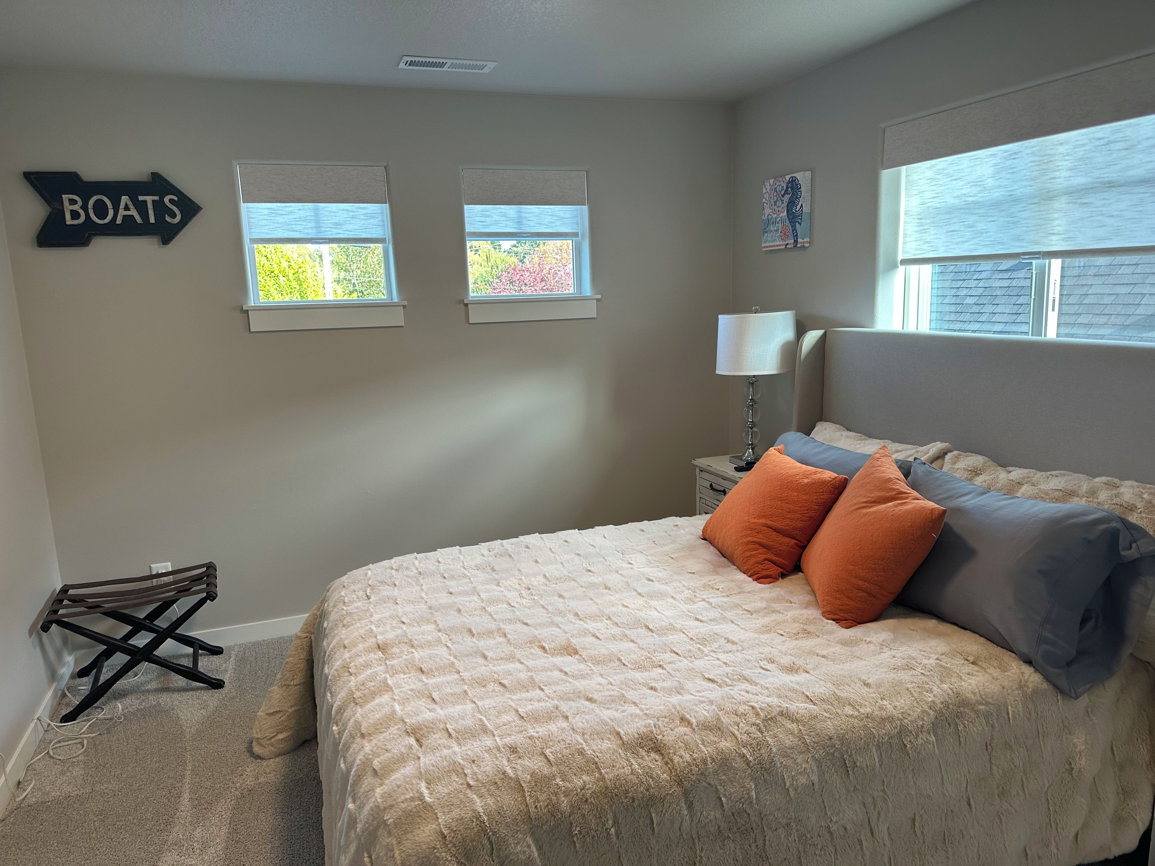 Un dormitorio con una cama, almohadas, una mesita de noche, una lámpara y dos ventanas.