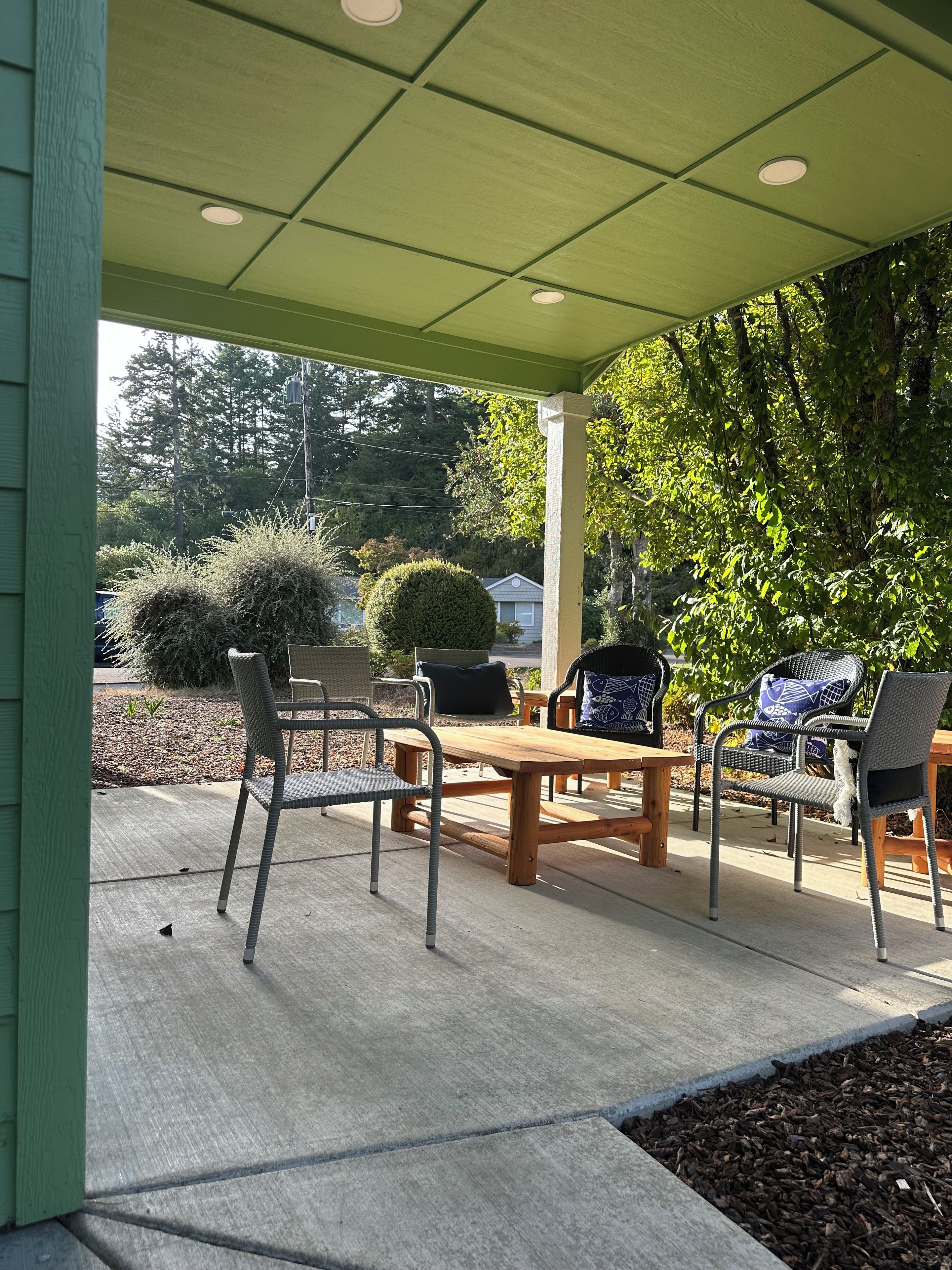 An outdoor patio with a wooden table and chairs, surrounded by greenery and a view of the neighborhood.