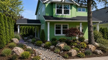 A two-story house with a green exterior, a stone pathway leading to the entrance, and a well-maintained garden with various plants and rocks.