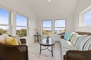 A bright room with a sofa, a table, and large windows offering a view of the outdoors. - East Sandwich House| Slps 8 | 3Beds2Baths (East Sandwich)