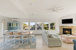 A spacious living room with a fireplace, a dining area with a table and chairs, and a large window offering a view of a house. - East Sandwich House| Slps 8 | 3Beds2Baths (East Sandwich)