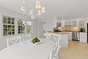 A modern kitchen with white cabinetry, a central island, and a dining area with a white table and chairs. - East Sandwich House| Slps 8 | 3Beds2Baths (East Sandwich)