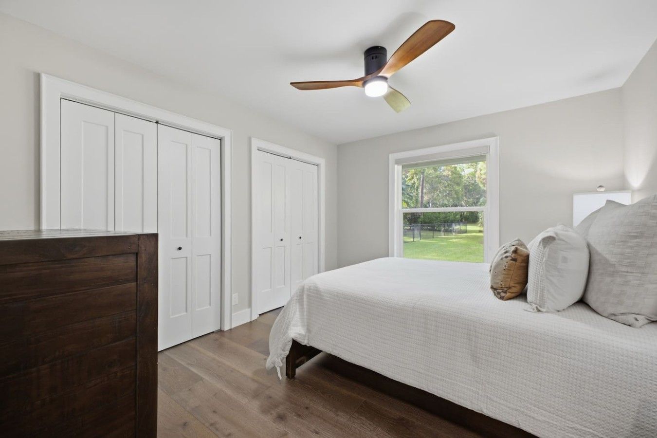 A bedroom with a bed, a ceiling fan, a window with a view of a green landscape, and a wooden dresser.