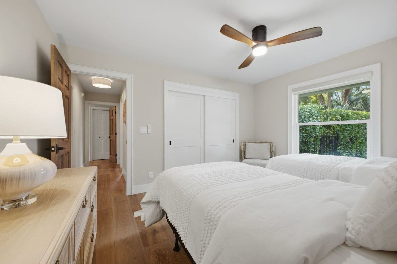 A bedroom with two beds, a ceiling fan, a nightstand with a lamp, and a window with a view of greenery.