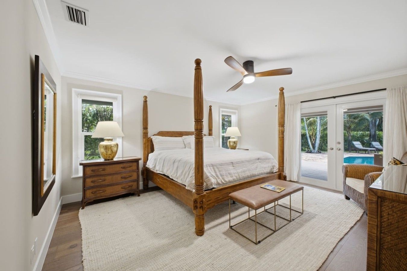 A bedroom with a four-poster bed, a ceiling fan, a nightstand with lamps, a bench, and a view of a swimming pool.