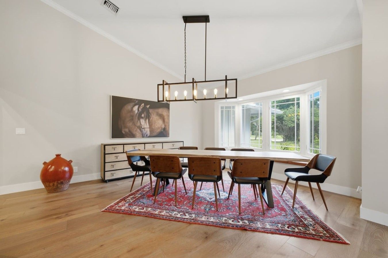 A dining area with a long table, six chairs, a red vase, a painting, and a large window with a view of greenery.