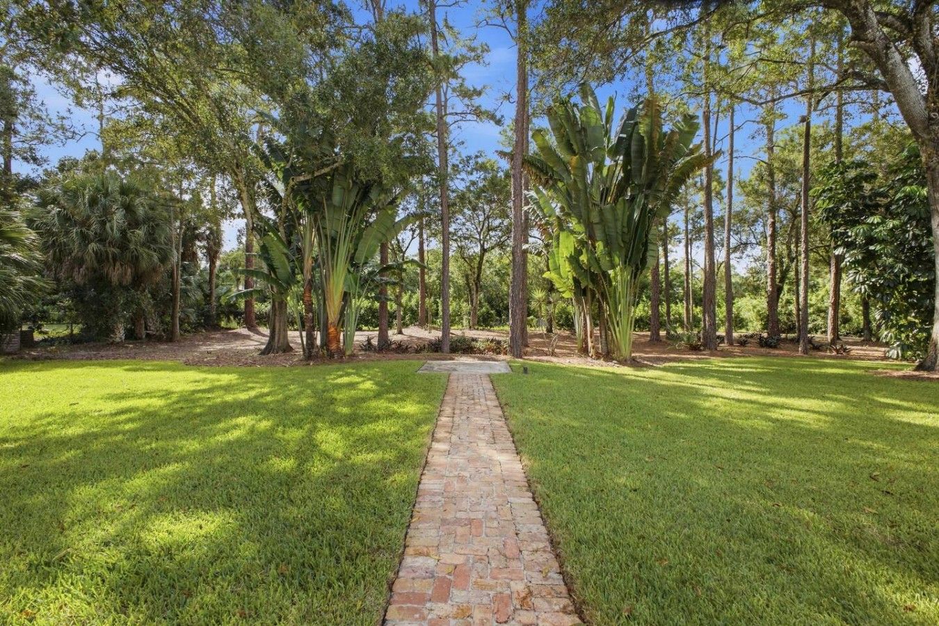 A paved pathway leading through a well-maintained grassy area with tall trees and a clear sky.